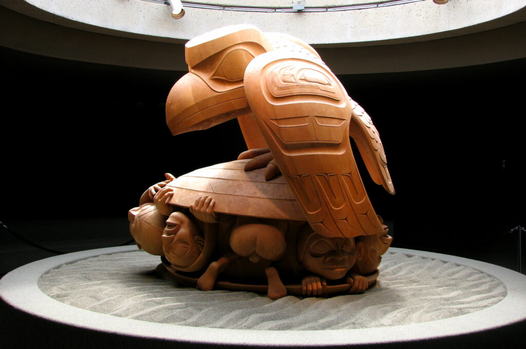An image of a piece of native artwork, a carved figure of a raven perched atop a clam from which humans emerge.
